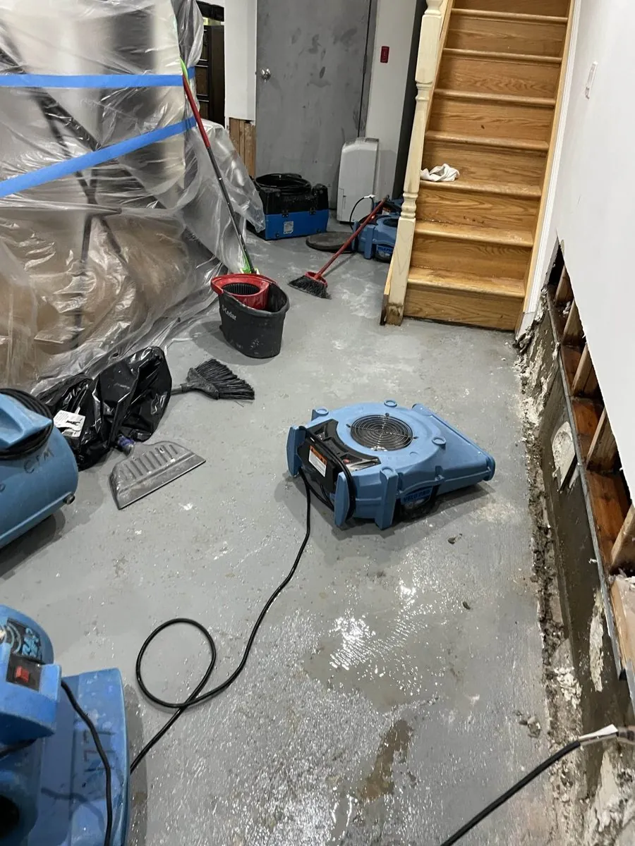 Air mover drying a flooded basement hallway with standing water near the stairs in Nassau County